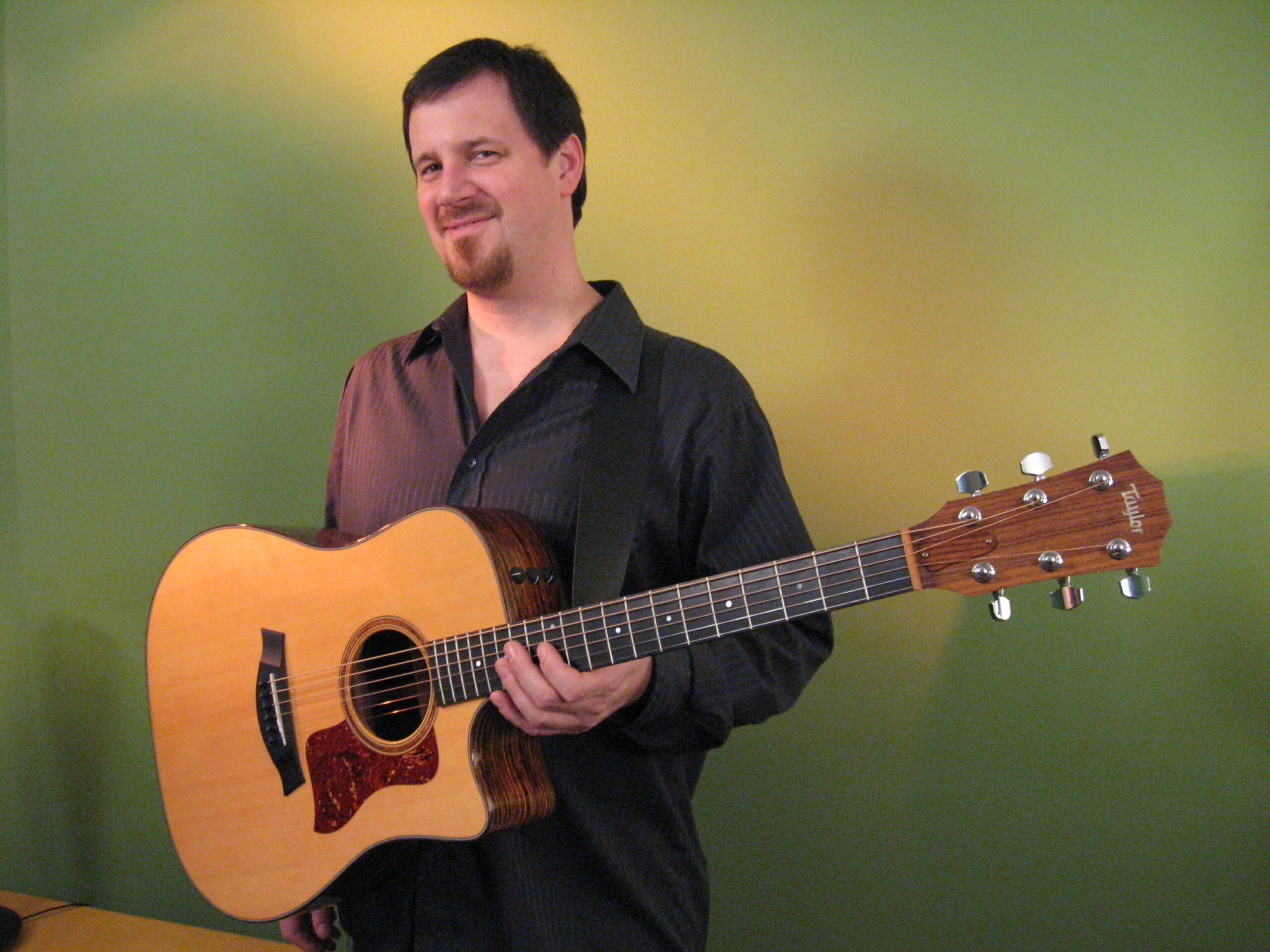 An adult holding an acoustic guitar while standing in front of a gradient green-yellow background