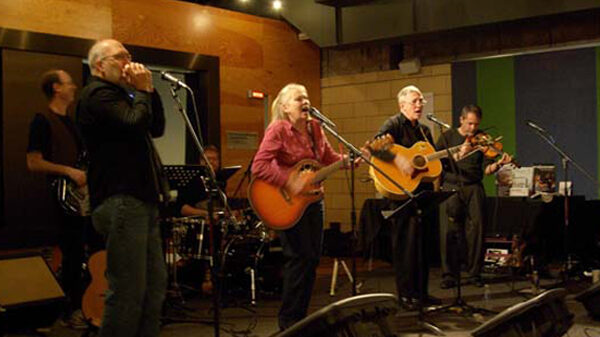 newlanders1 An adult playing a harmonica, an adult playing an electric bass, two adults playing acoustic guitars and singing into microphones, and an adult playing a violin in front of a radio studio