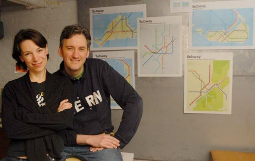Two adults posing next to maps on a gray wall