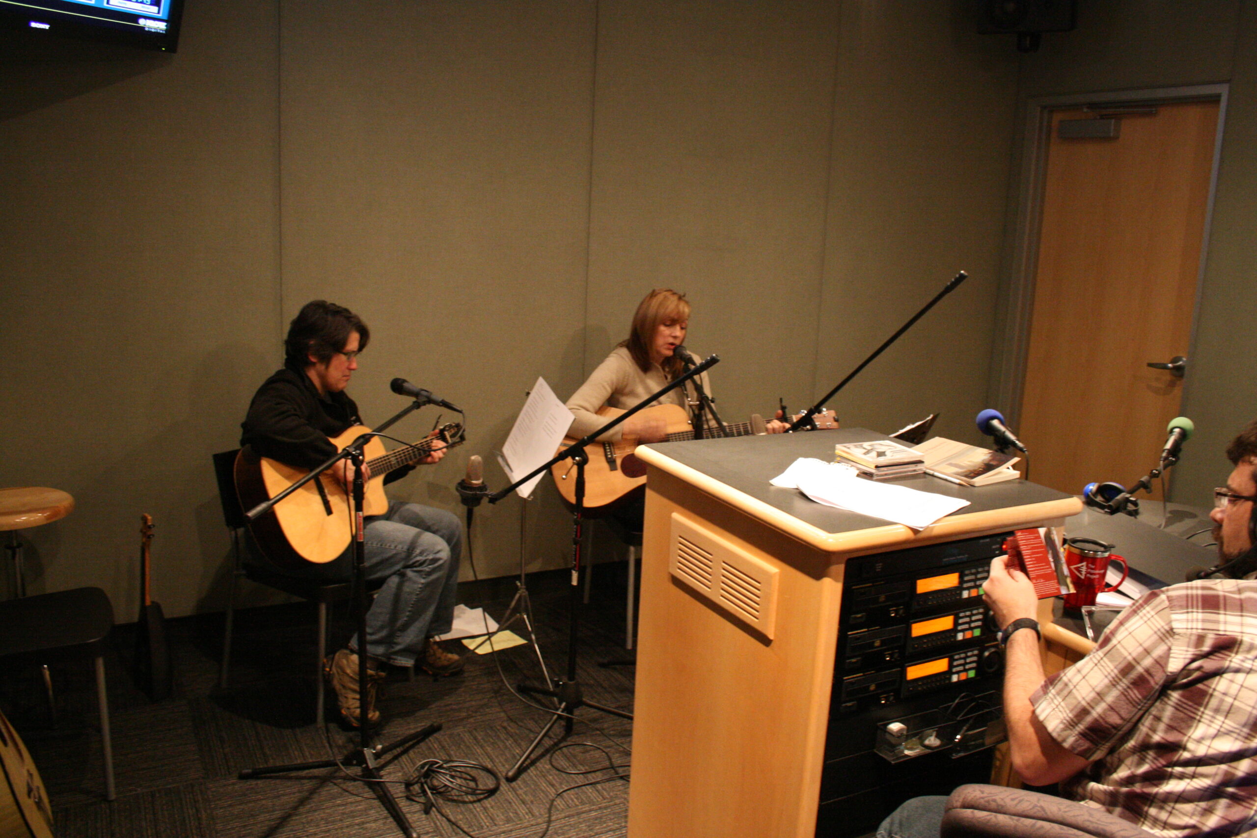 Two adults playing acoustic guitars and singing into microphones in a radio studio