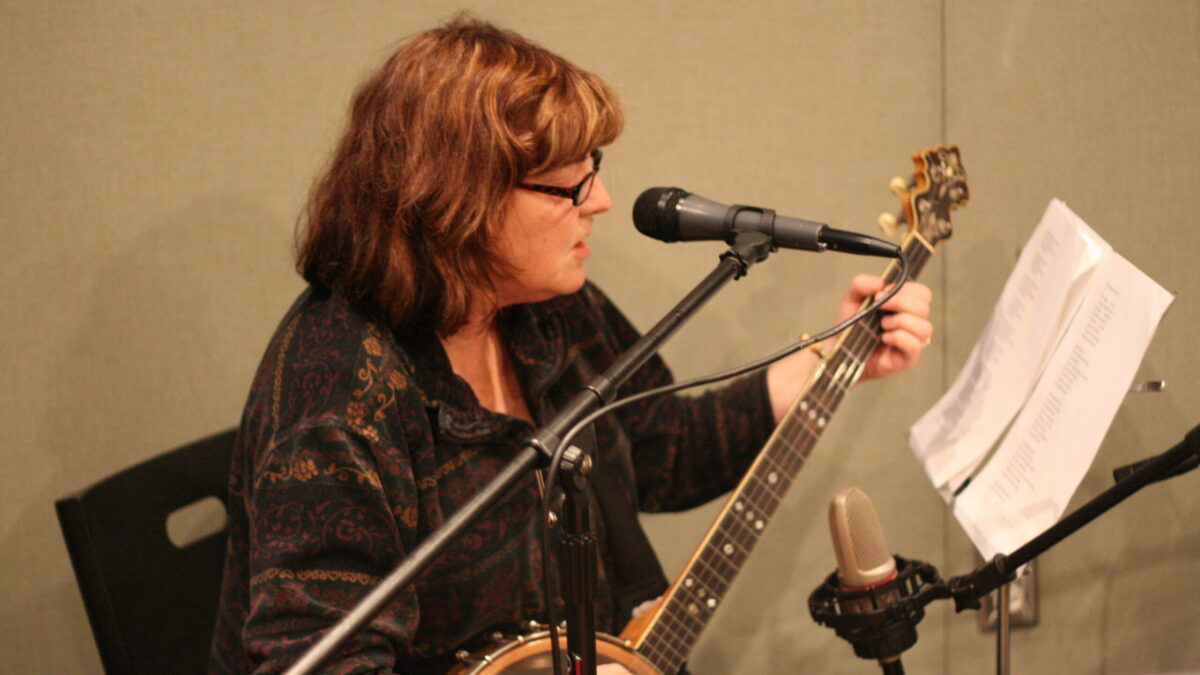 Sue Powers An adult playing an acoustic guitar and singing into a microphone in a radio studio
