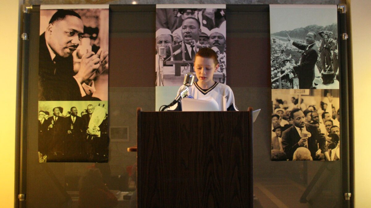 A youth standing behind a podium speaking into a microphone in front of banners of Martin Luther King, Jr.