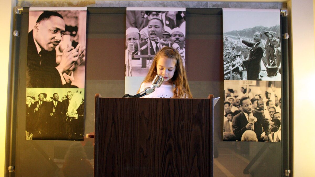 A youth standing behind a podium speaking into a microphone in front of banners of Martin Luther King, Jr.