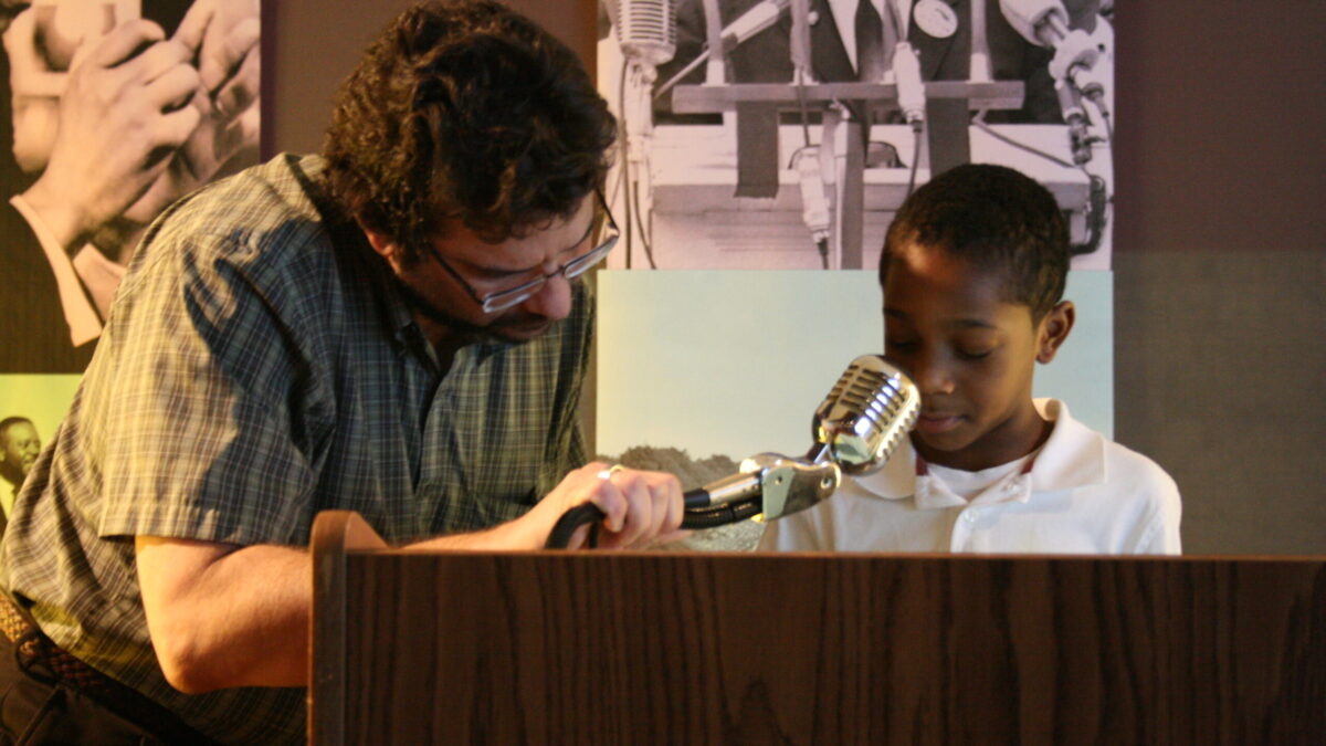 A youth and an adult standing behind a podium speaking into a microphone in front of banners of Martin Luther King, Jr.