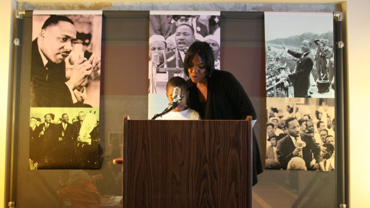 A youth and an adult standing behind a podium speaking into a microphone in front of banners of Martin Luther King, Jr.
