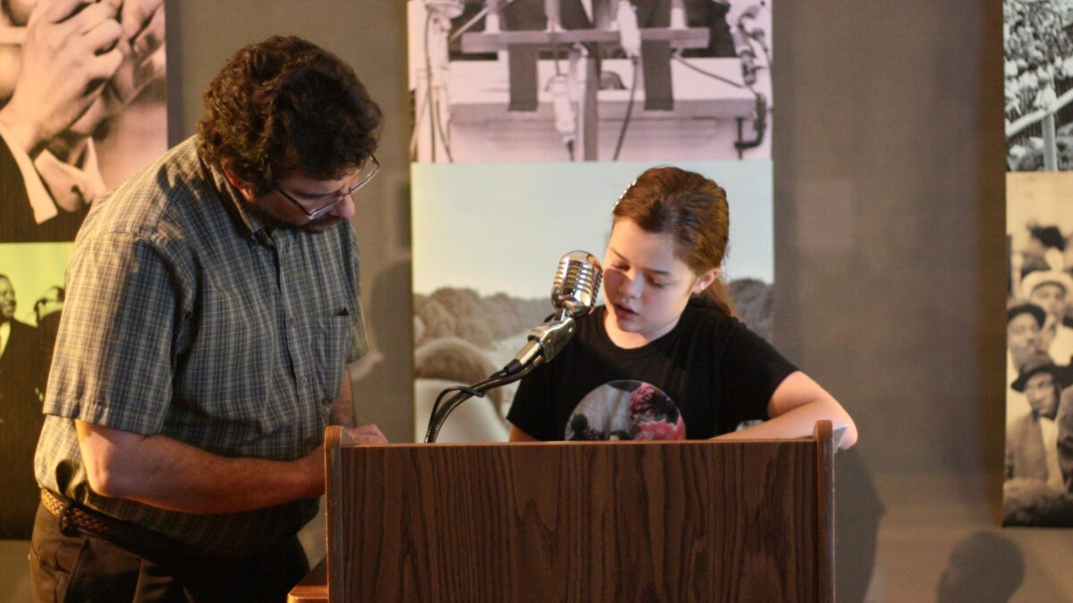 A youth and an adult standing behind a podium speaking into a microphone in front of banners of Martin Luther King, Jr.