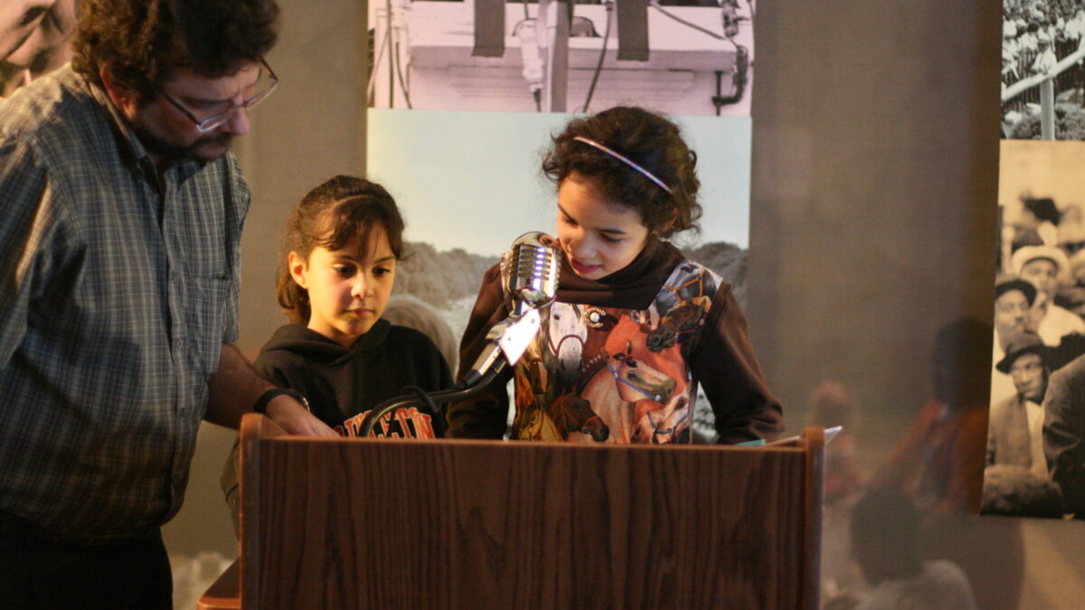 Two youth and an adult standing behind a podium speaking into a microphone in front of banners of Martin Luther King, Jr.