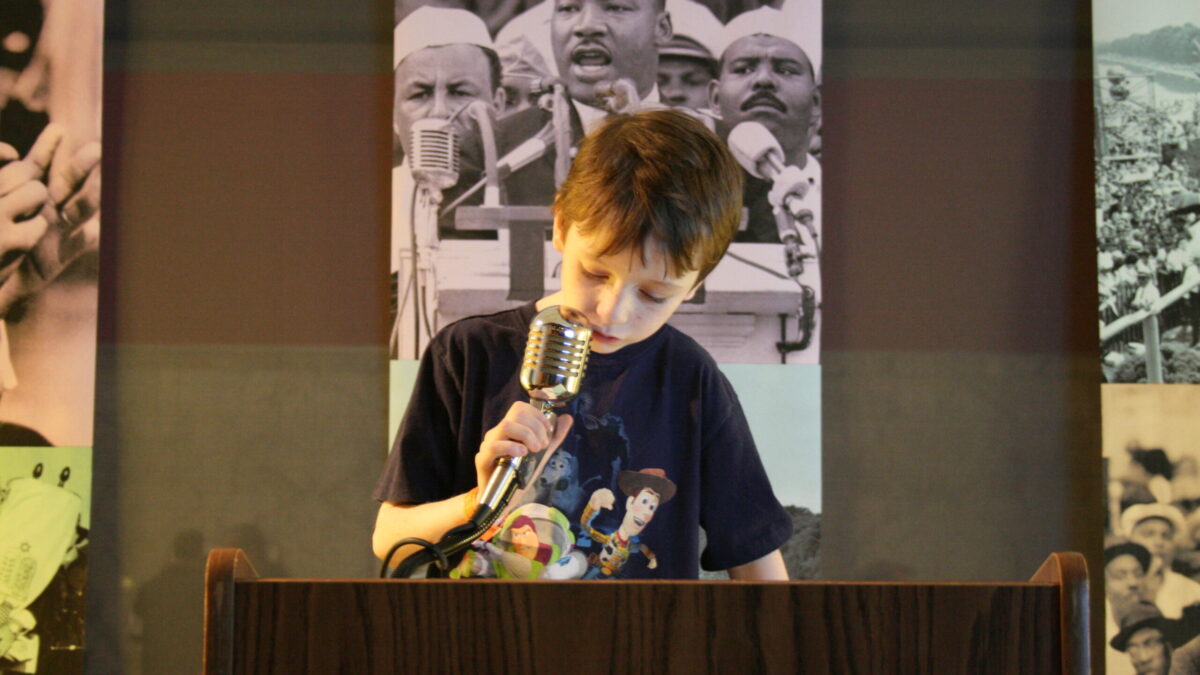 A youth standing behind a podium speaking into a microphone in front of banners of Martin Luther King, Jr.