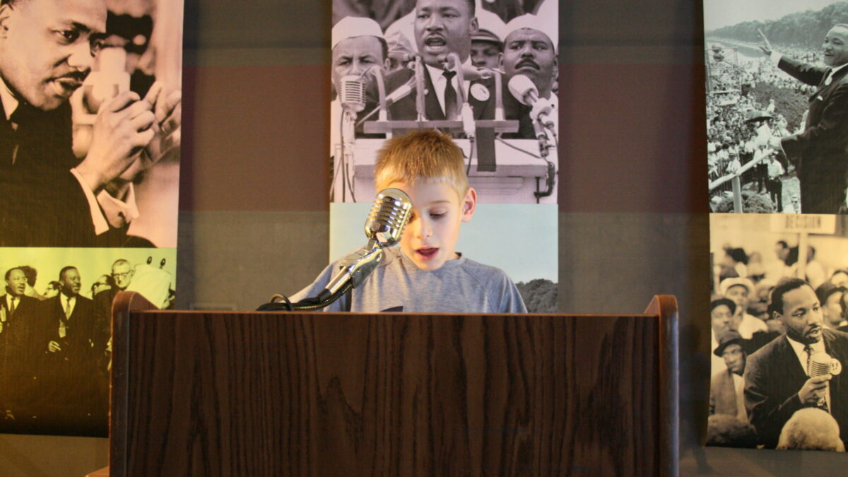 A youth standing behind a podium speaking into a microphone in front of banners of Martin Luther King, Jr.