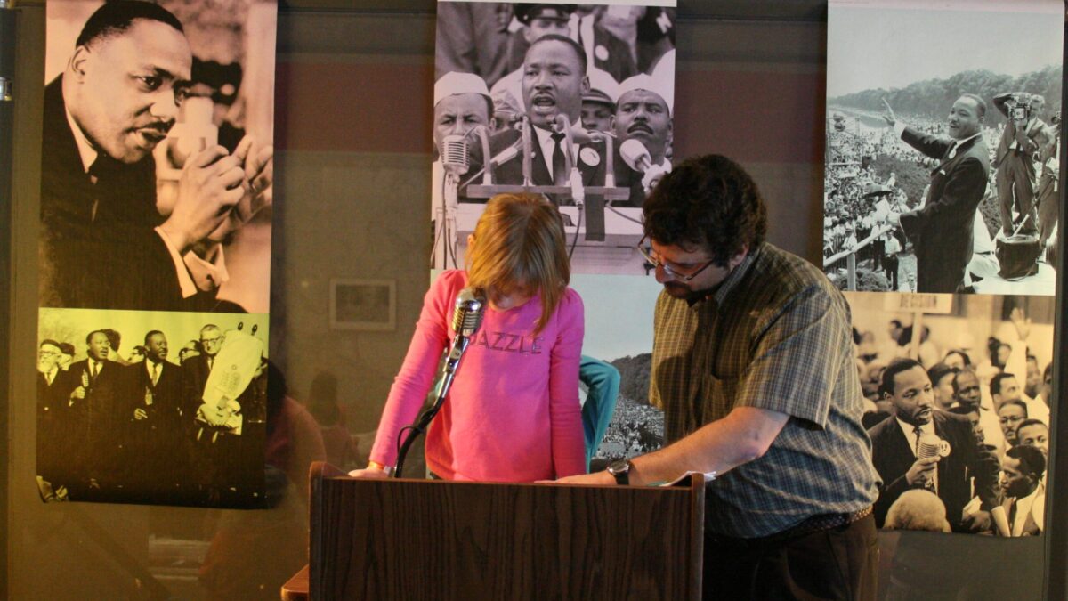 A youth and an adult standing behind a podium speaking into a microphone in front of banners of Martin Luther King, Jr.