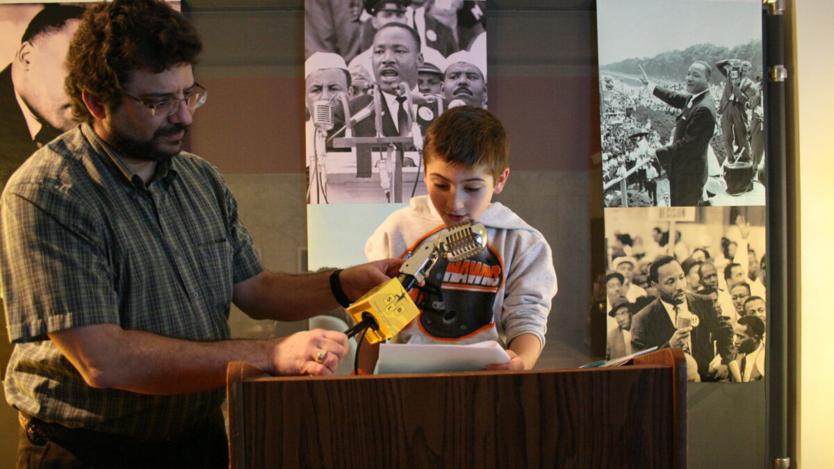 A youth and an adult standing behind a podium speaking into a microphone in front of banners of Martin Luther King, Jr.