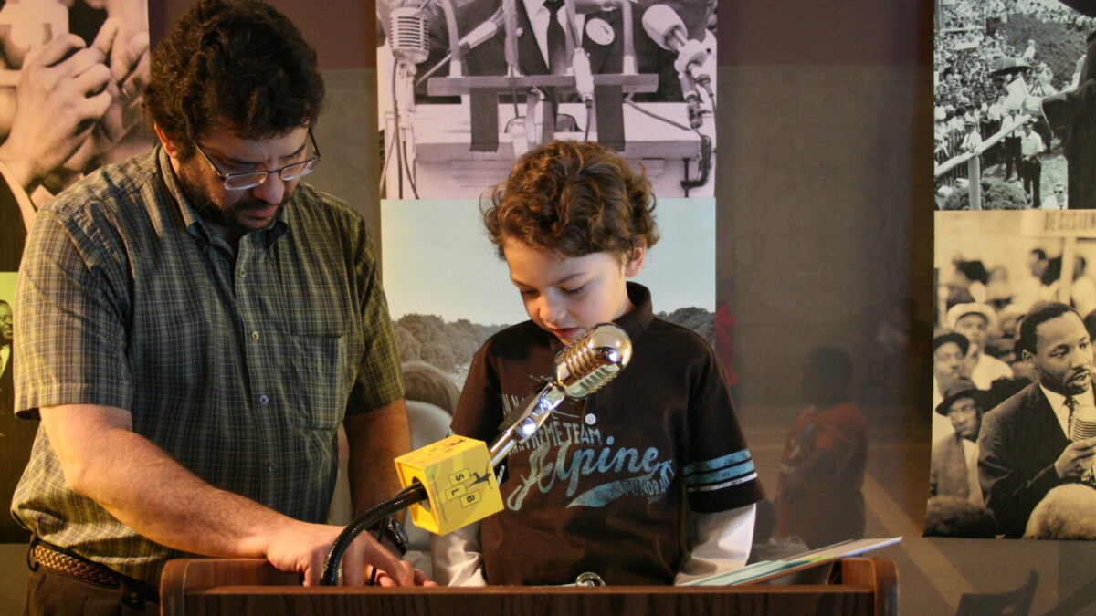 A youth and an adult standing behind a podium speaking into a microphone in front of banners of Martin Luther King, Jr.