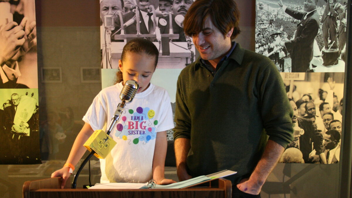 A youth and an adult standing behind a podium speaking into a microphone in front of banners of Martin Luther King, Jr.