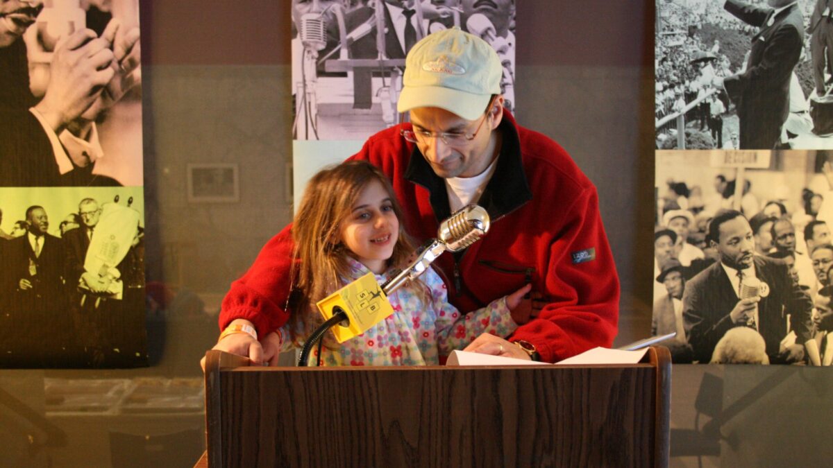 A youth and an adult standing behind a podium speaking into a microphone in front of banners of Martin Luther King, Jr.