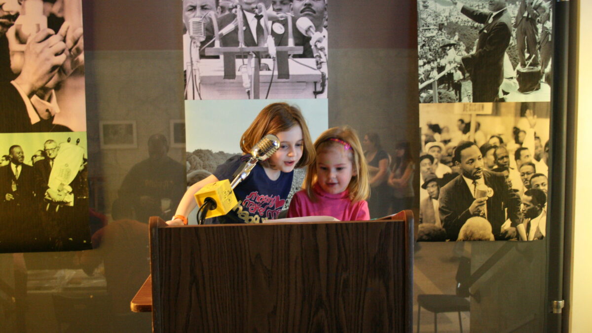 Two youth standing behind a podium speaking into a microphone in front of banners of Martin Luther King, Jr.