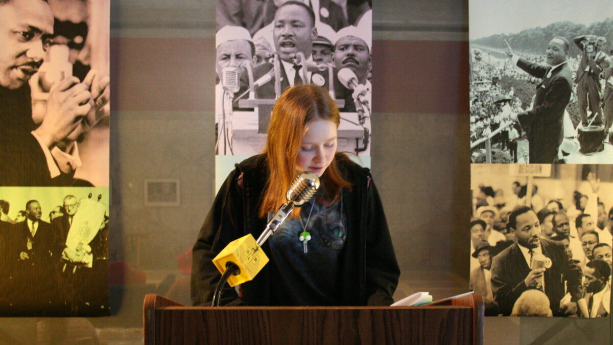 A youth standing behind a podium speaking into a microphone in front of banners of Martin Luther King, Jr.