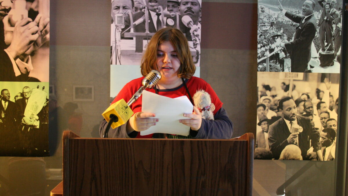 A youth standing behind a podium speaking into a microphone in front of banners of Martin Luther King, Jr.