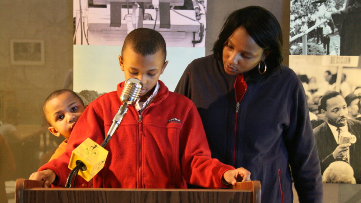 Two youth and an adult standing behind a podium speaking into a microphone in front of banners of Martin Luther King, Jr.