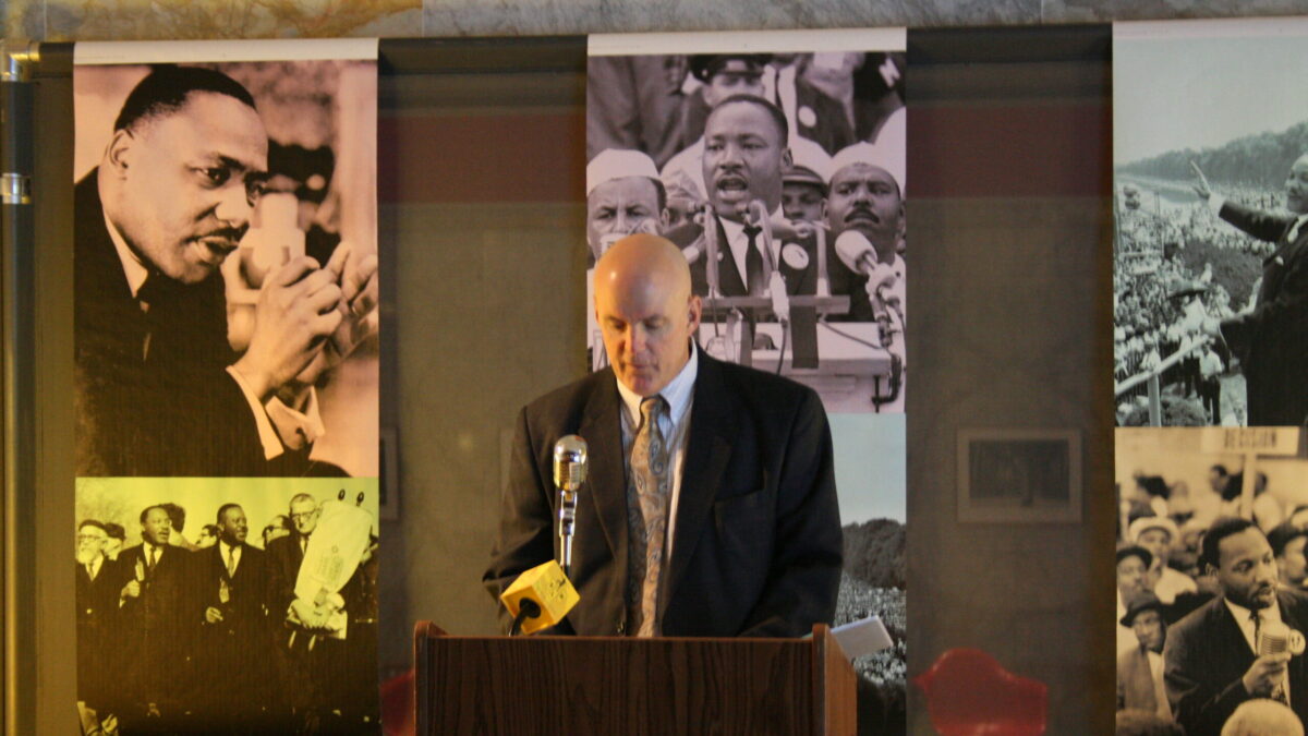 An adult standing behind a podium speaking into a microphone in front of banners of Martin Luther King, Jr.