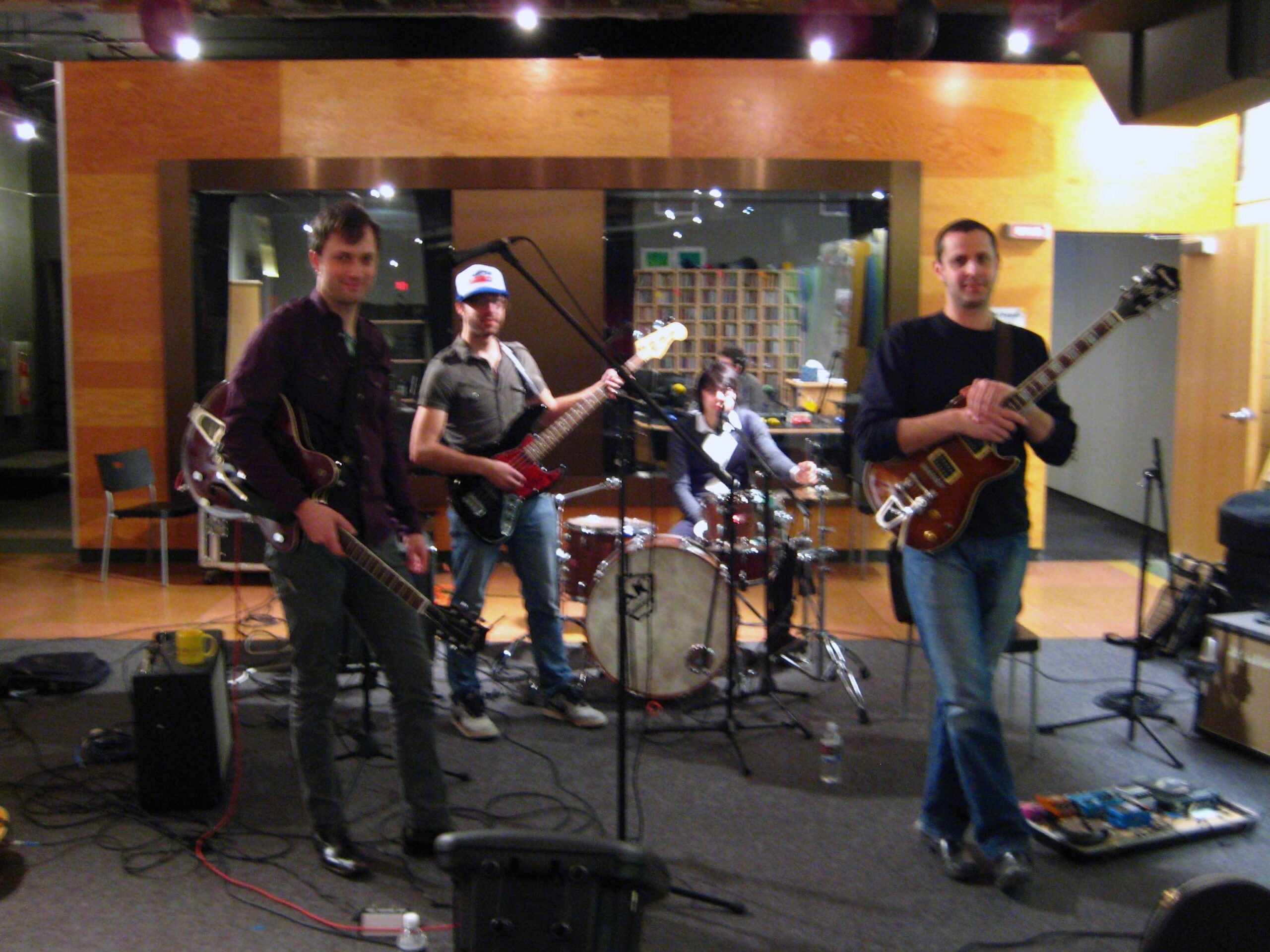 Two adults holding electric guitars, an adult holding an electric bass, and an adult sitting at a drum set in front of a radio studio