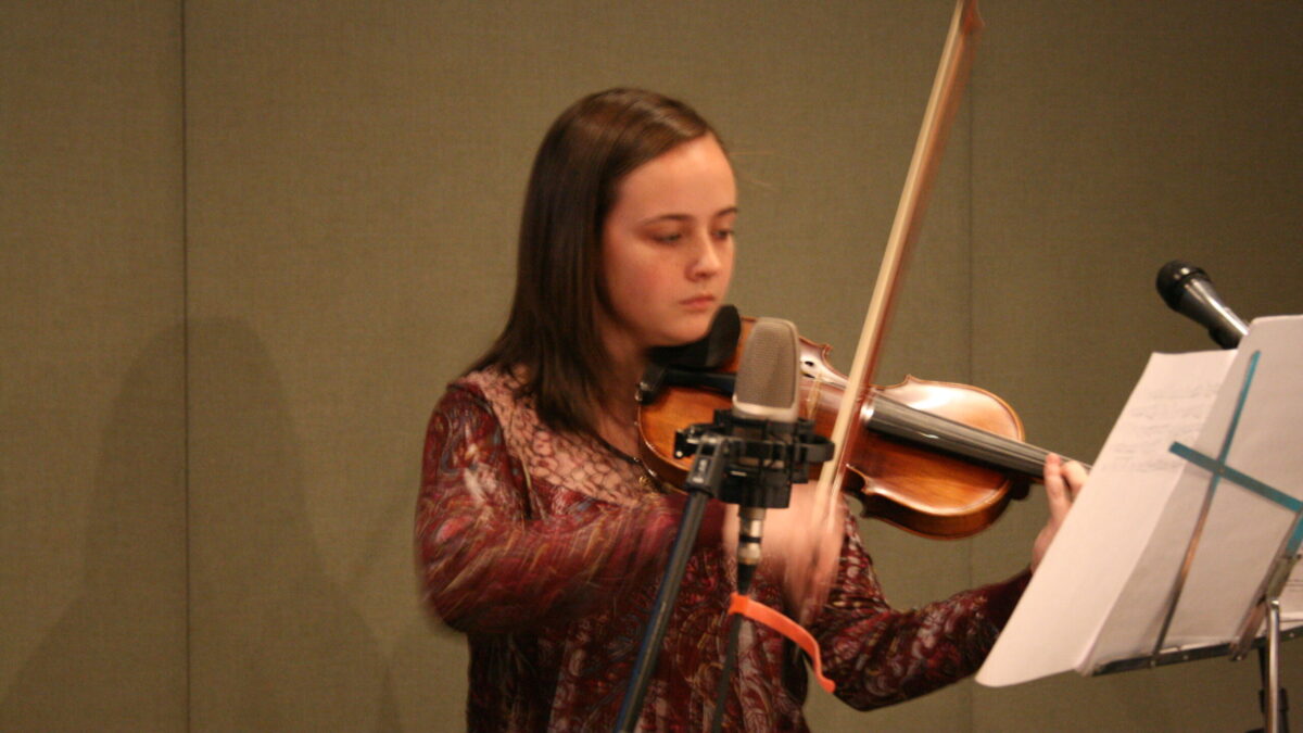A youth playing a violin and looking at sheet music on a music stand in a radio studio