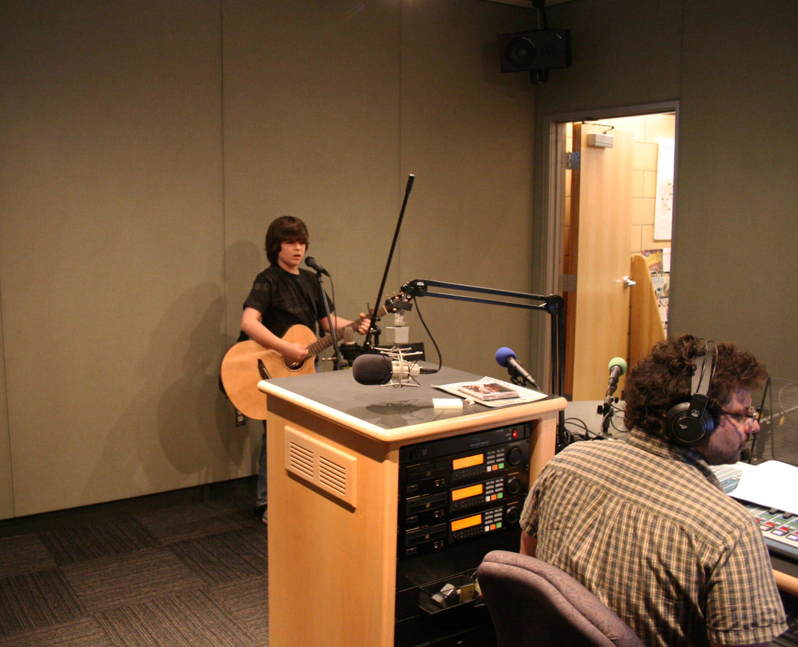 A teen holding an acoustic guitar and speaking into a microphone being interviewed by an adult wearing headphones in a radio studio