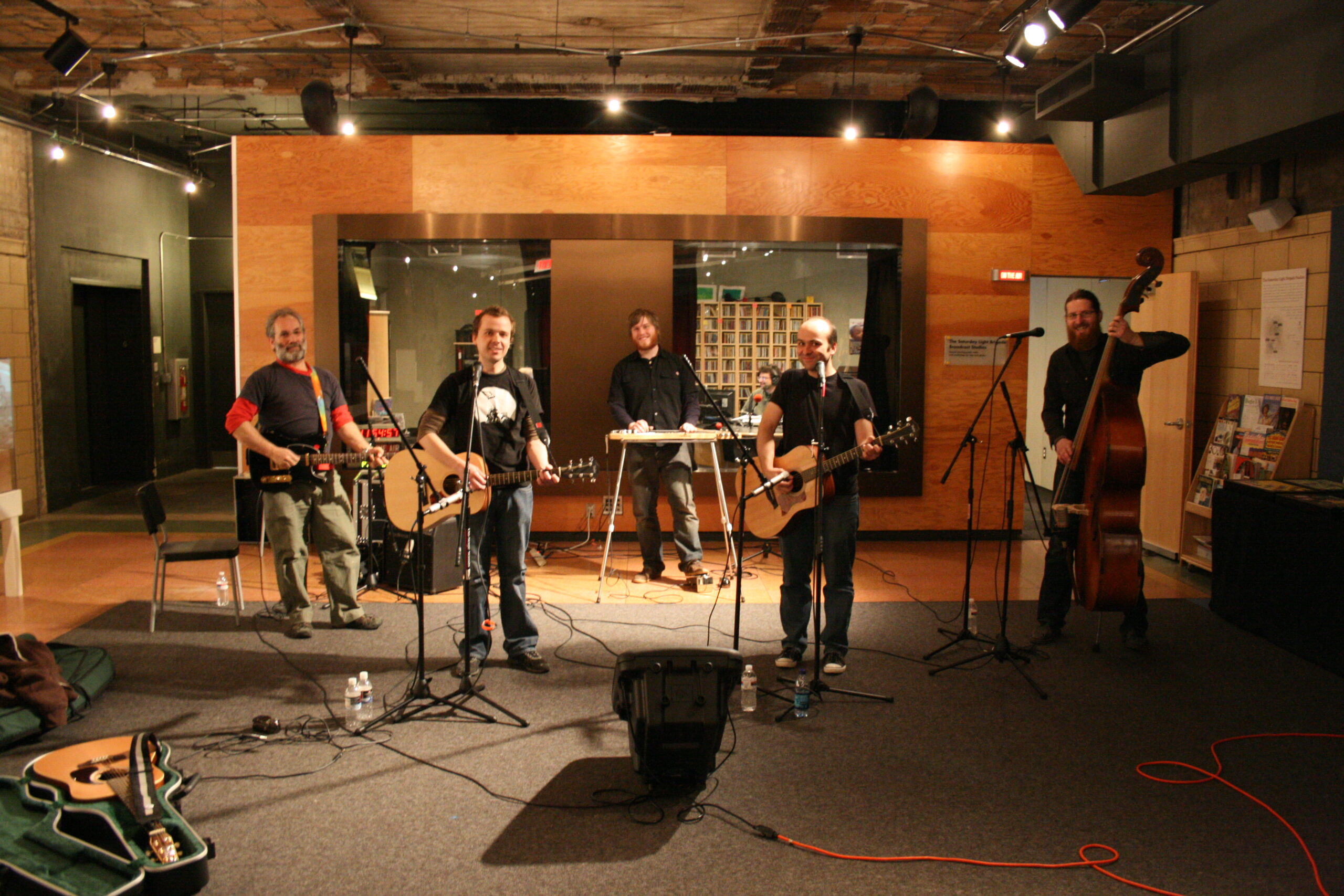 An adult playing electric guitar, two adults playing acoustic guitar, an adult playing a piano keyboard, and an adult playing an upright bass in front of a radio studio