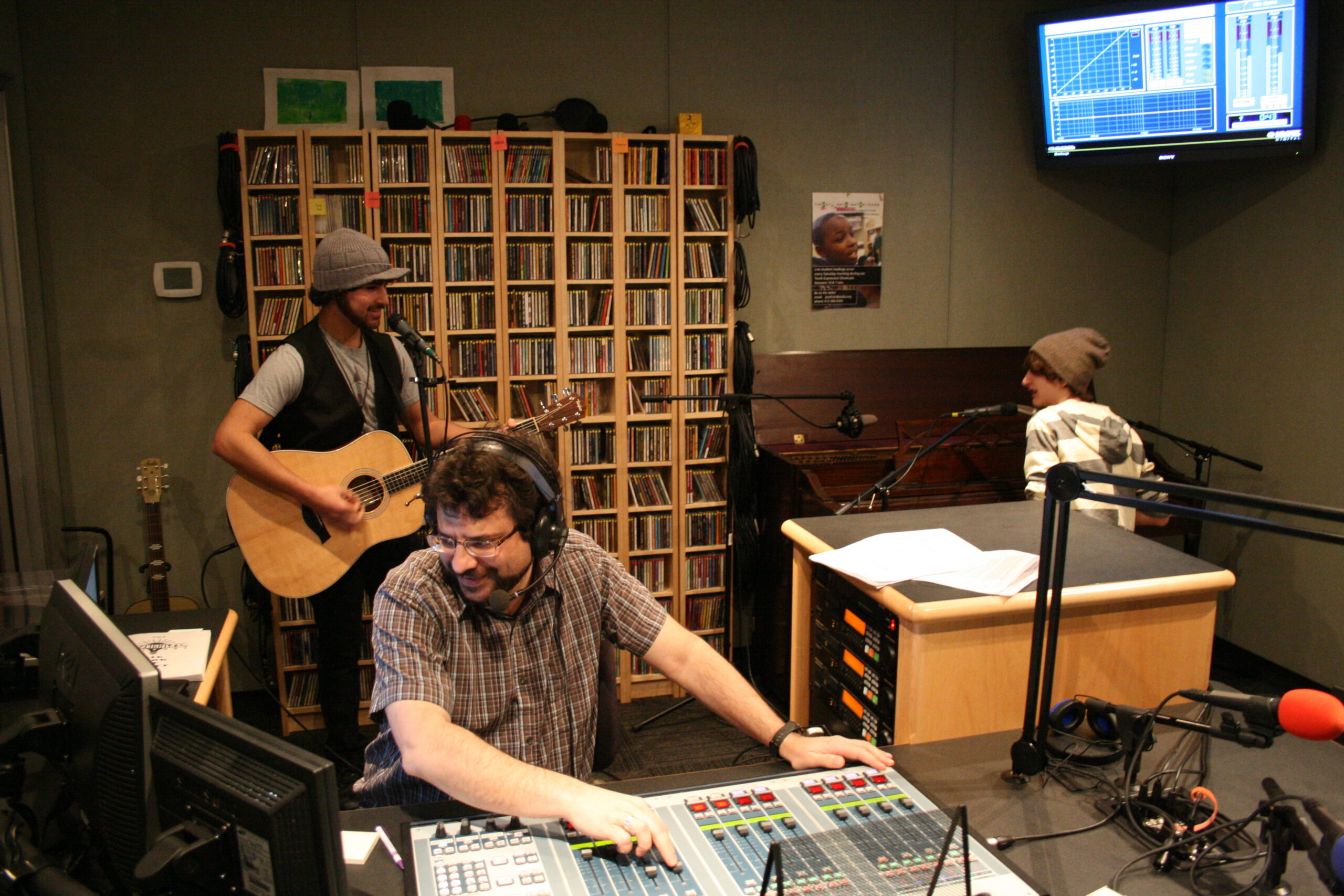 A teen playing acoustic guitar and a teen playing piano as an adult wearing headphones listens in a radio studio