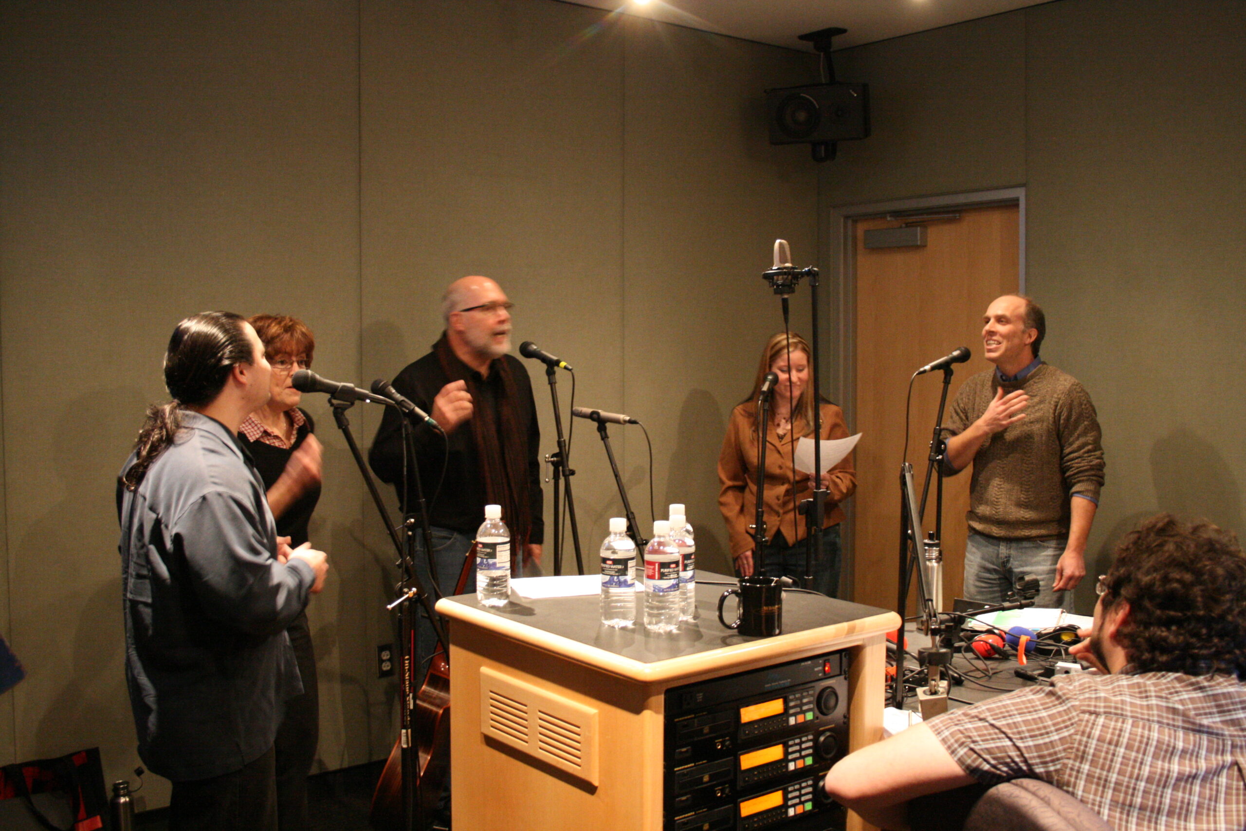 Five adults singing into microphones as an adult listens in a radio studio