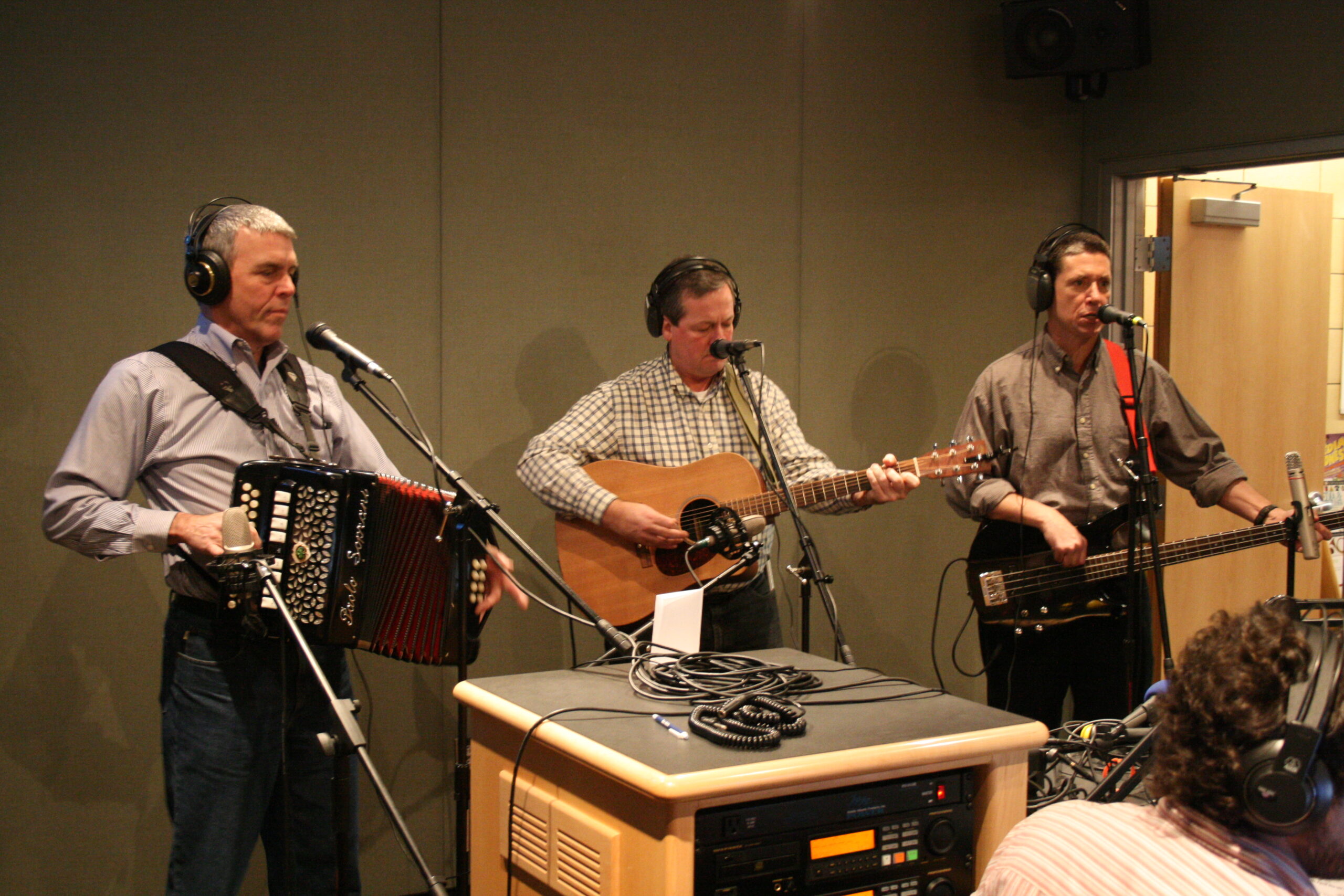 An adult wearing headphones playing an accordion, an adult playing acoustic guitar, and an adult playing electric guitar in a radio studio