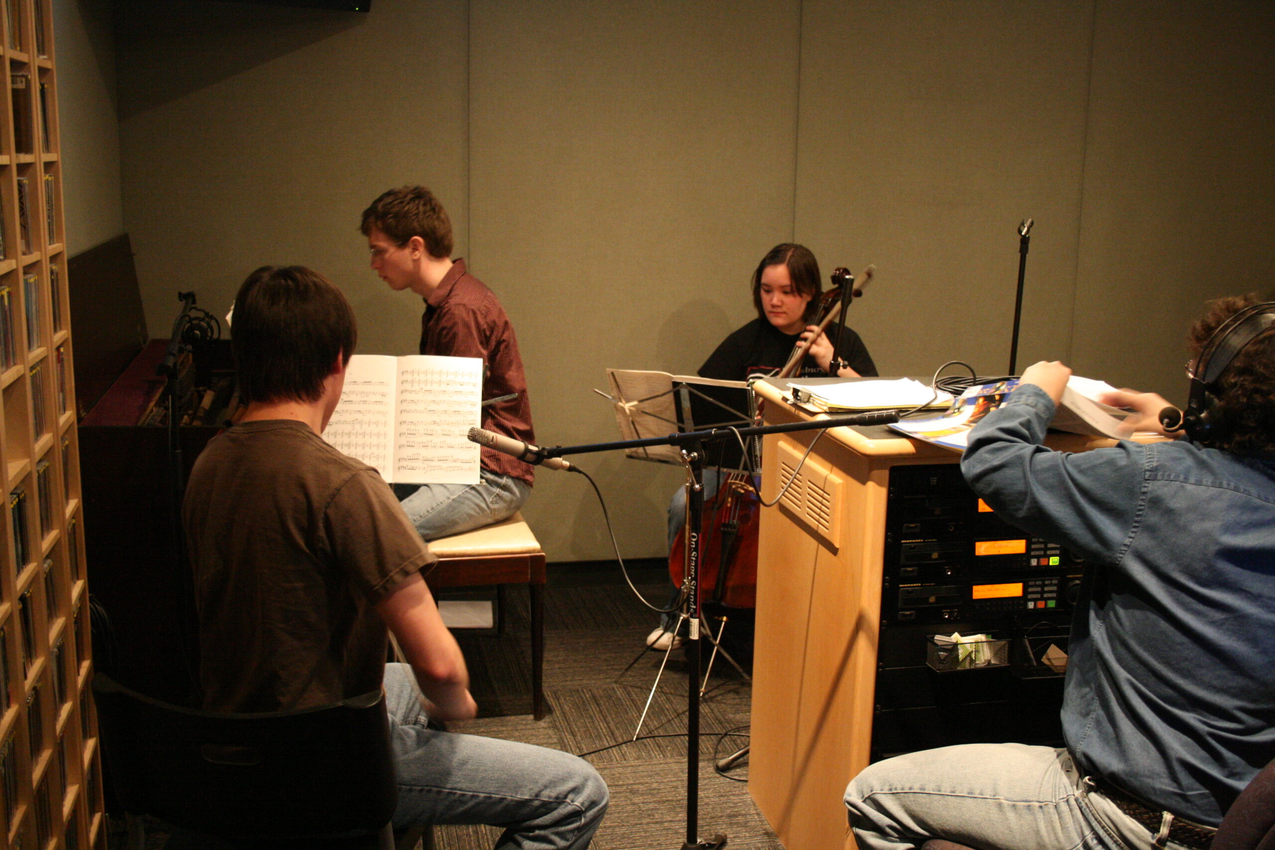 A teen playing a piano, a teen sitting behind a music stand, and a teen playing a cello looking at sheet music on a music stand as an adult listens in a radio studio