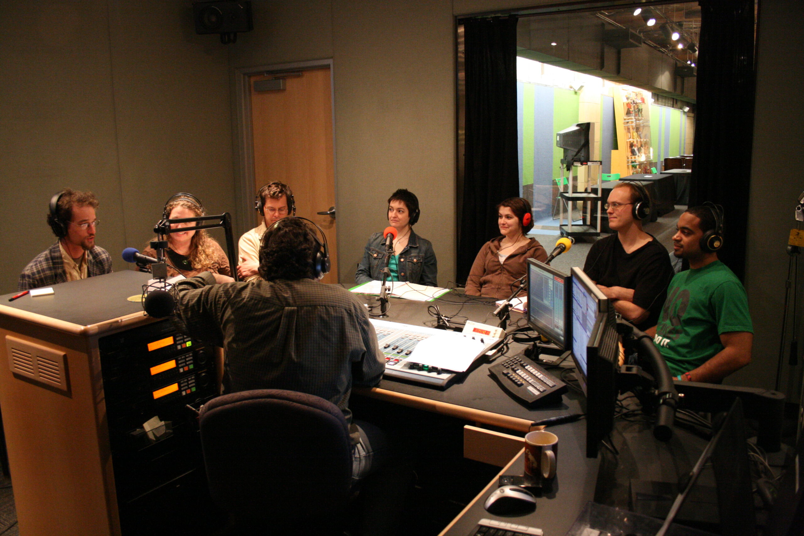 Seven adults sitting behind microphones being interviewed by an adult wearing headphones sitting behind an audio mixer in a radio studio
