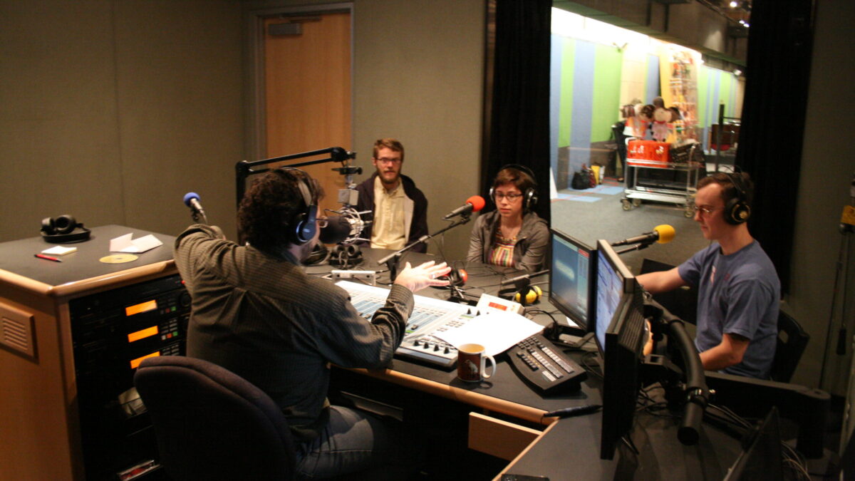Two adults and a teen wearing headphones speaking into microphones being interviewed by an adult wearing headphones sitting behind an audio mixer in a radio studio