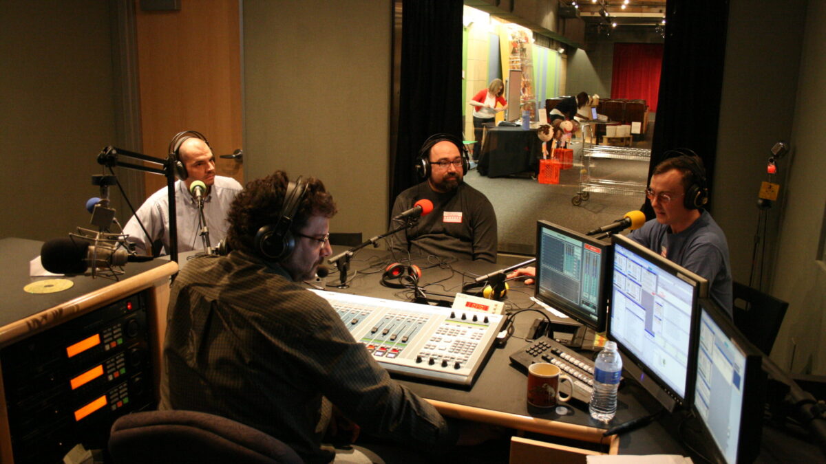 Three adults wearing headphones speaking into microphones being interviewed by an adult wearing headphones sitting behind an audio mixer in a radio studio