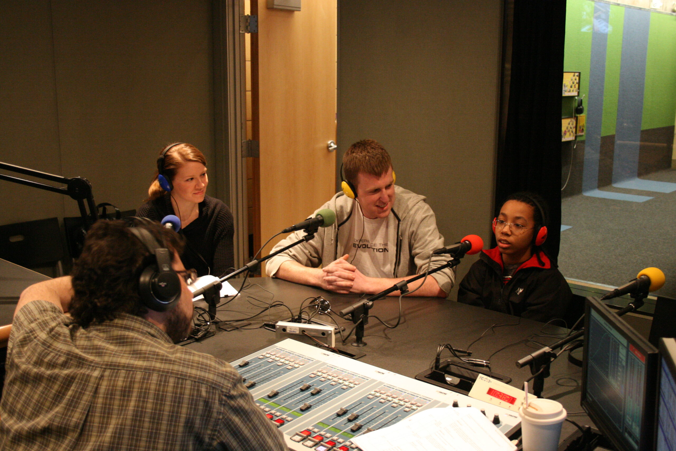 Two adults and a youth wearing headphones speaking into microphones being interviewed by an adult wearing headphones and sitting behind an audio mixer in a radio studio