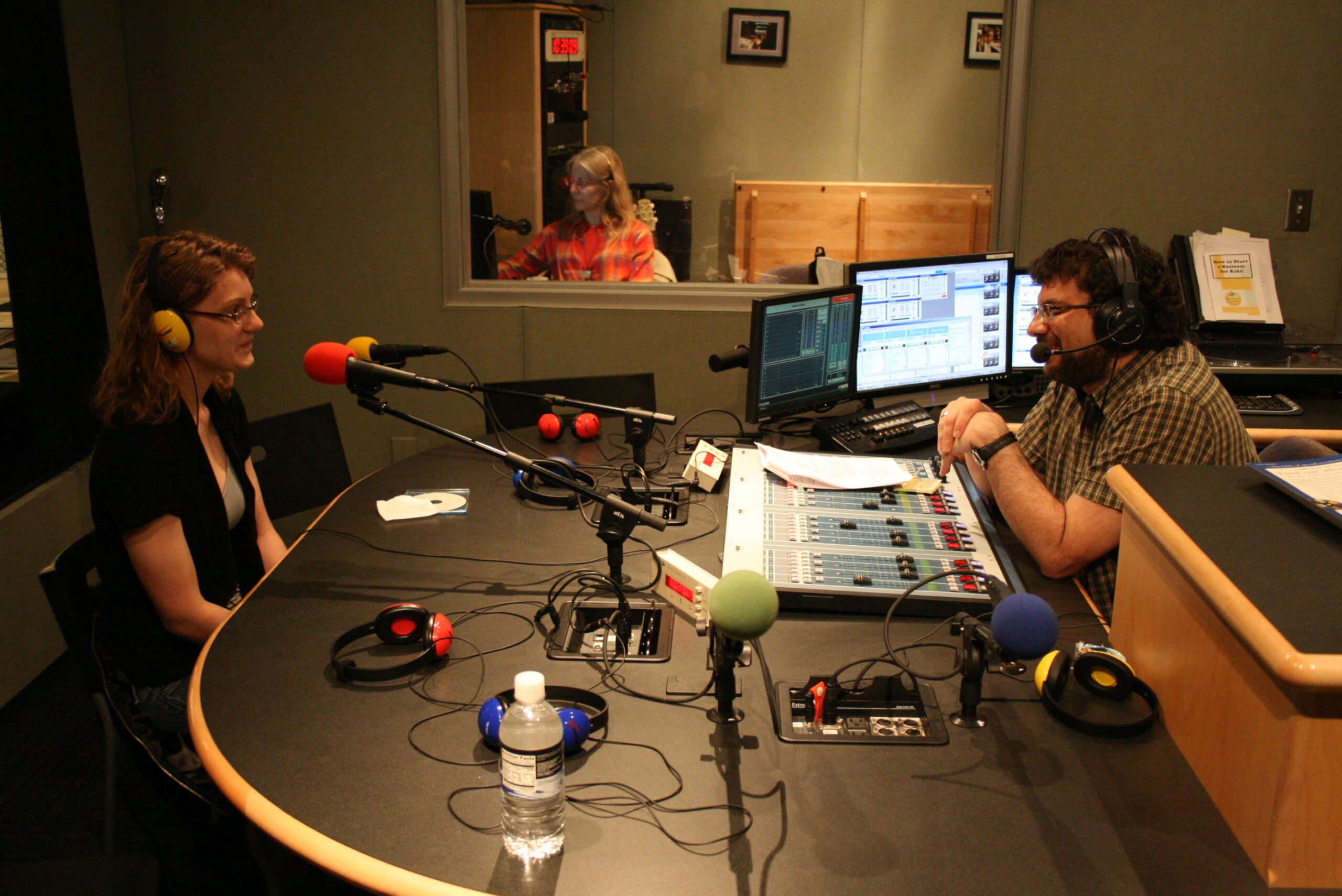 A teen wearing headphones sitting behind a microphone being interviewed by an adult wearing headphones sitting behind an audio mixer in a radio studio