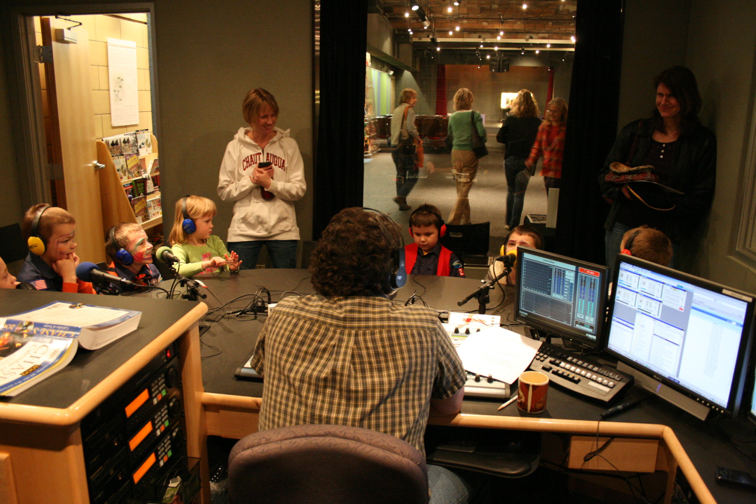 Seven youth wearing headphones sitting behind microphones being interviewed by an adult as two adults stand in the background in a radio studio