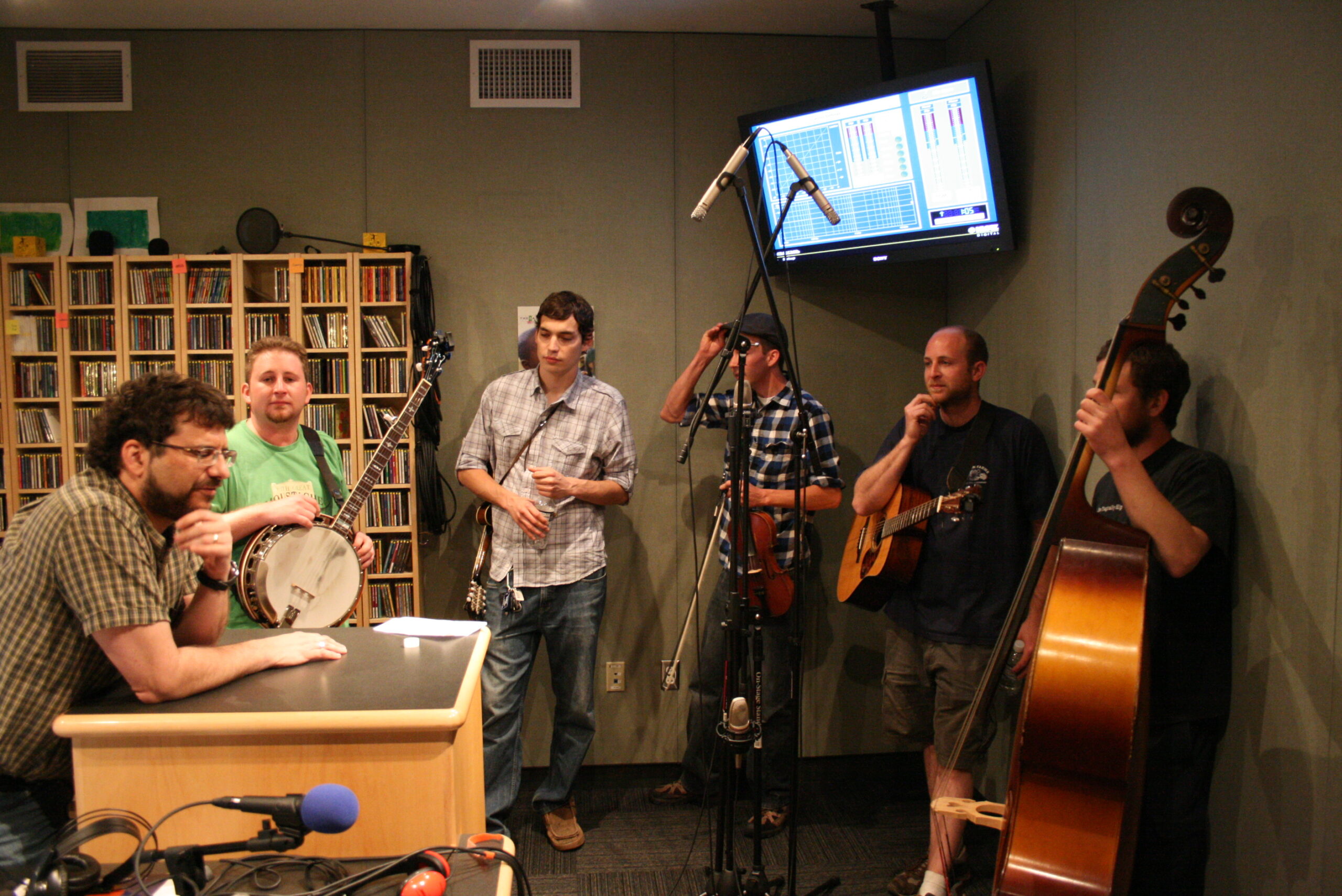 An adult interviewing an adult holding a banjo, an adult holding a mandolin, an adult holding a violin, an adult holding an acoustic guitar, and an adult holding an upright bass in a radio studio