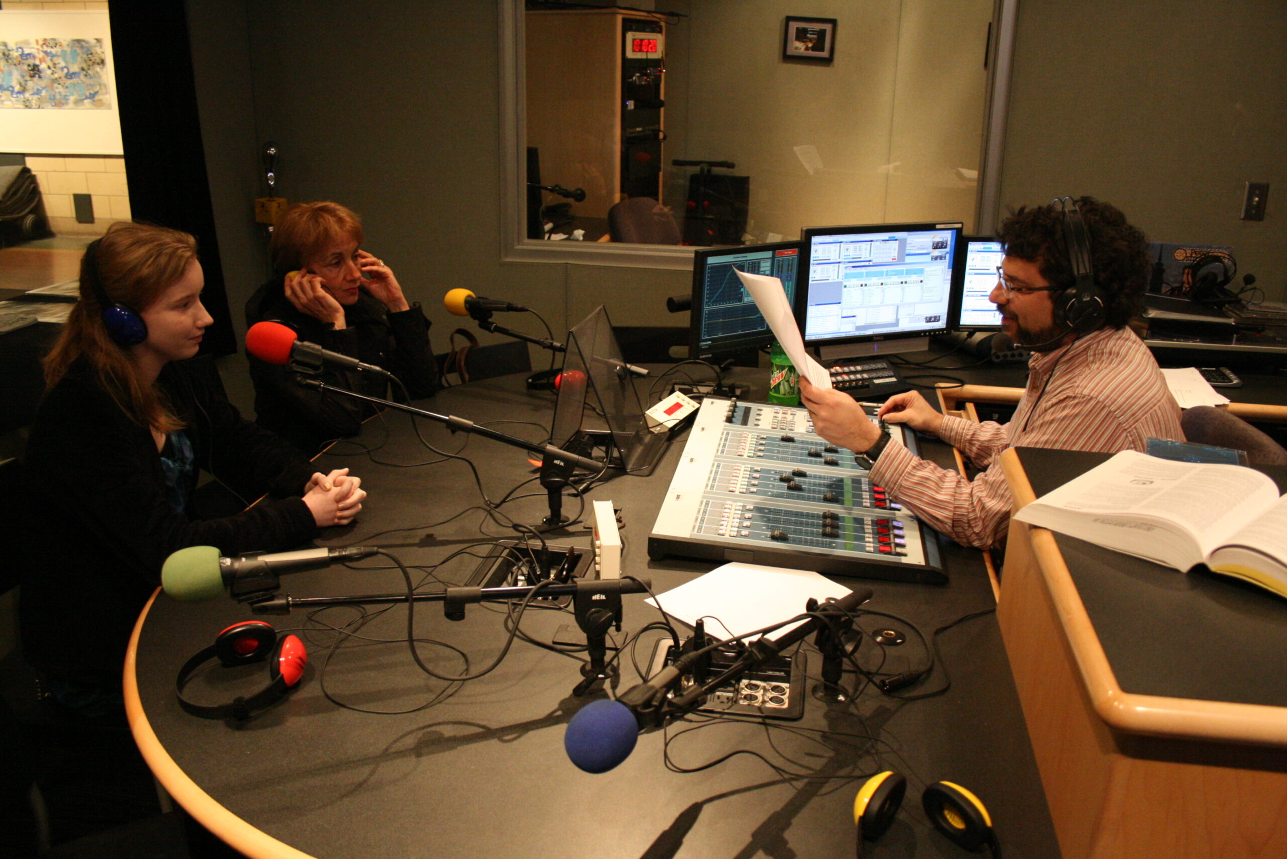 A teen and an adult wearing headphones and speaking into microphones being interviewed by an adult sitting behind an audio mixer while holding a piece of paper in a radio studio
