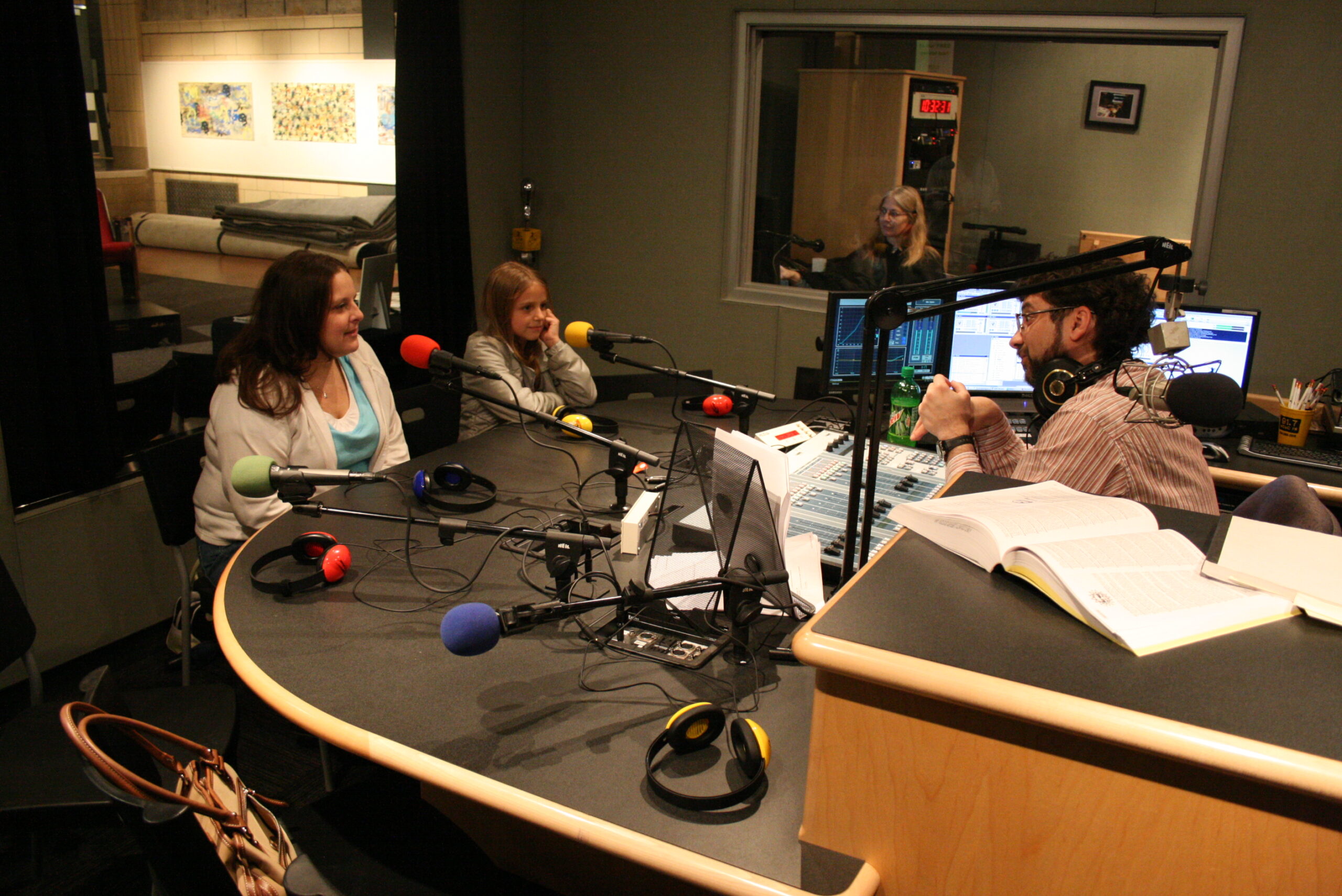 An adult and a youth sitting behind microphones being interviewed by an adult sitting behind a microphone and an audio mixer in a radio studio