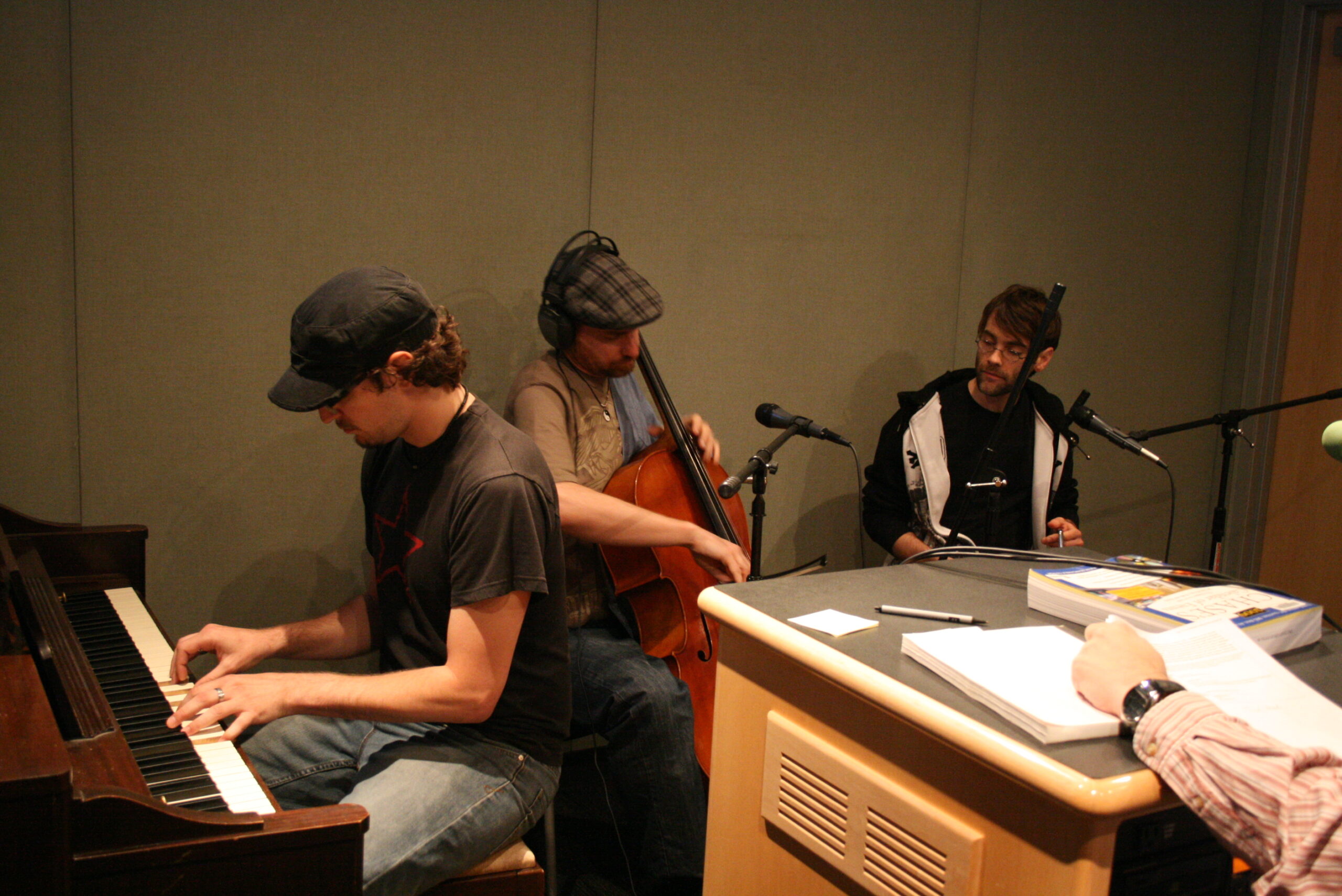 An adult playing a piano, an adult wearing headphones playing a cello, and an adult sitting behind a microphone in a radio studio