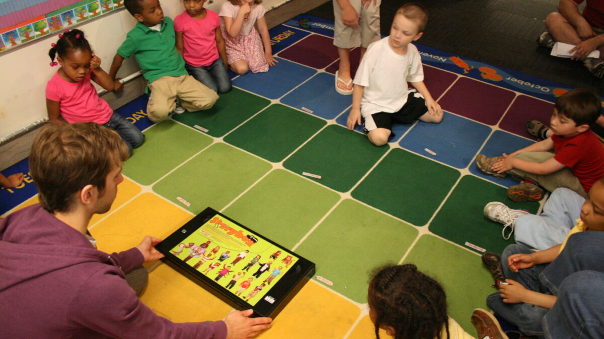 Nine youth and an adult sitting around a tablet on a multicolored classroom floor
