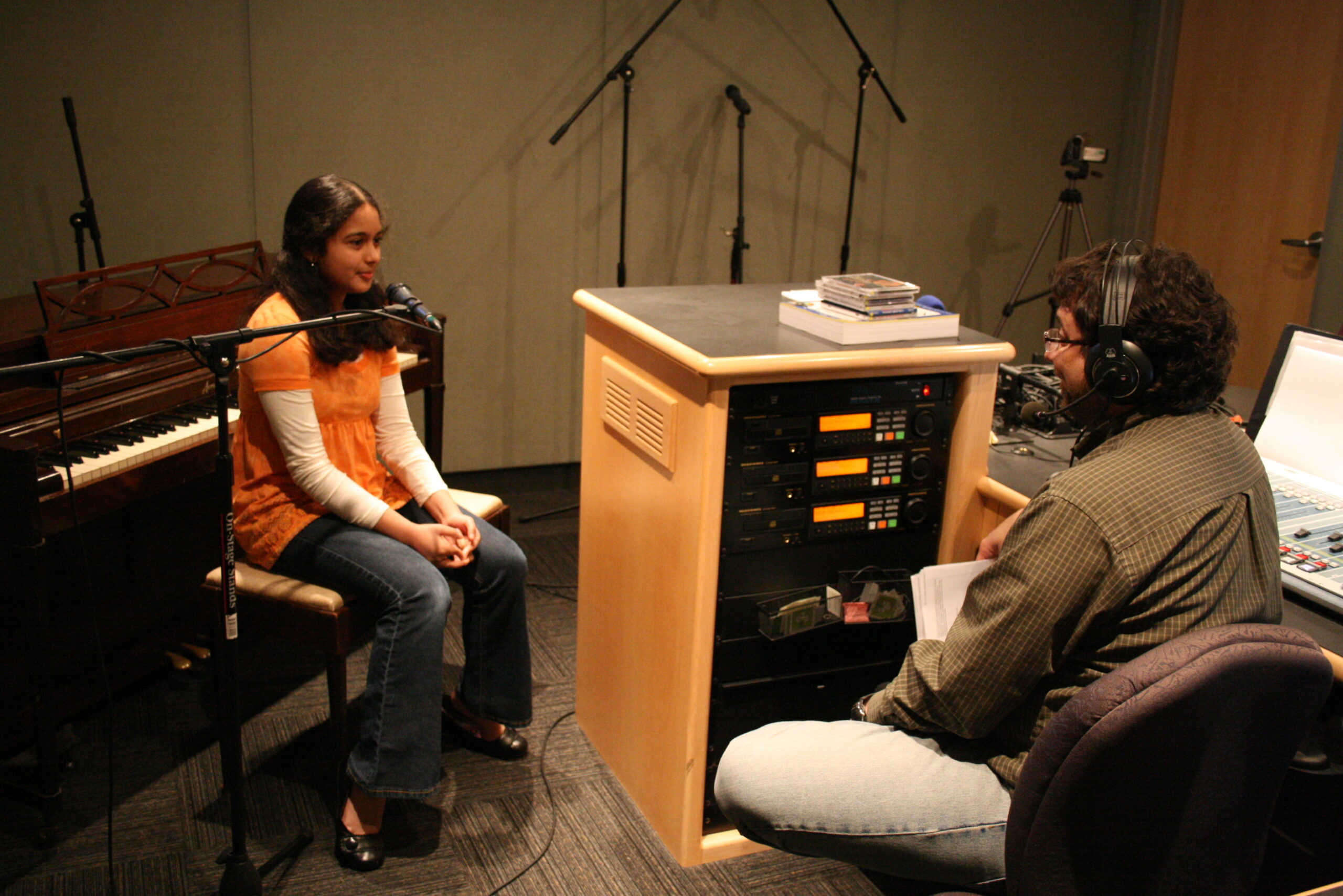 A teen sitting at a piano being interviewed by an adult wearing headphones in a radio studio