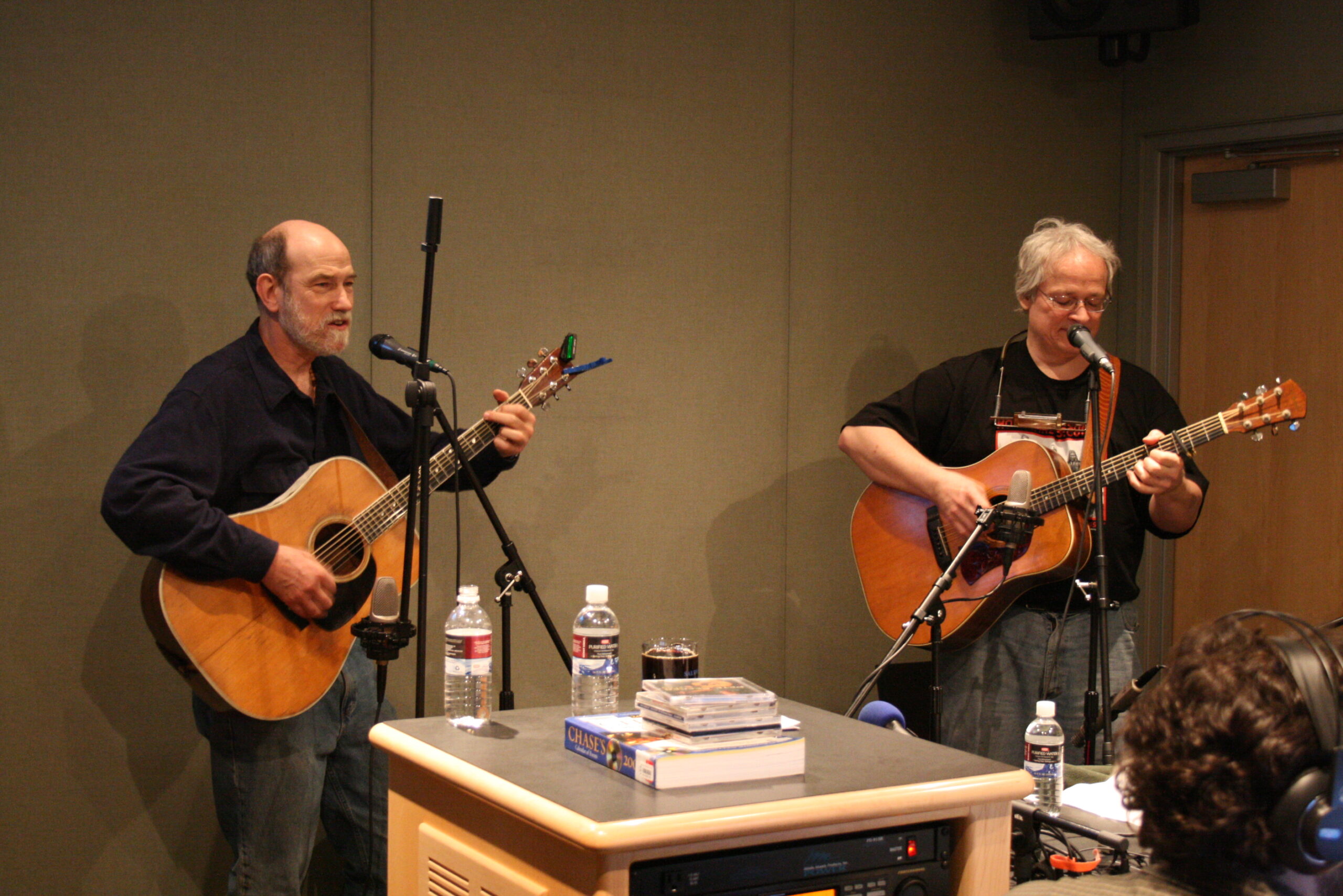 Two adults playing acoustic guitars and singing into microphones in a radio studio