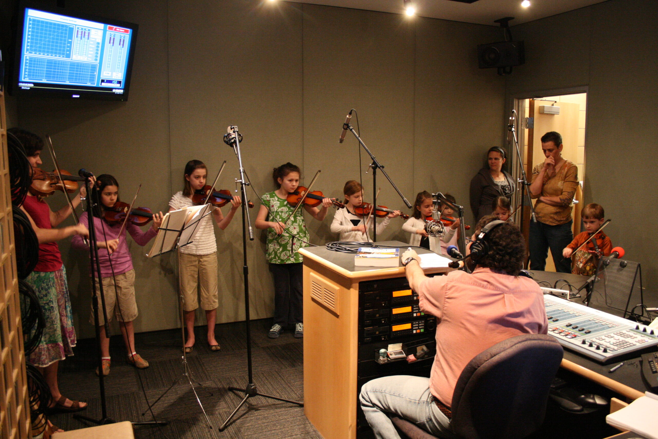 Eight youth playing violins as three adults, one wearing headphones, listens in a radio studio