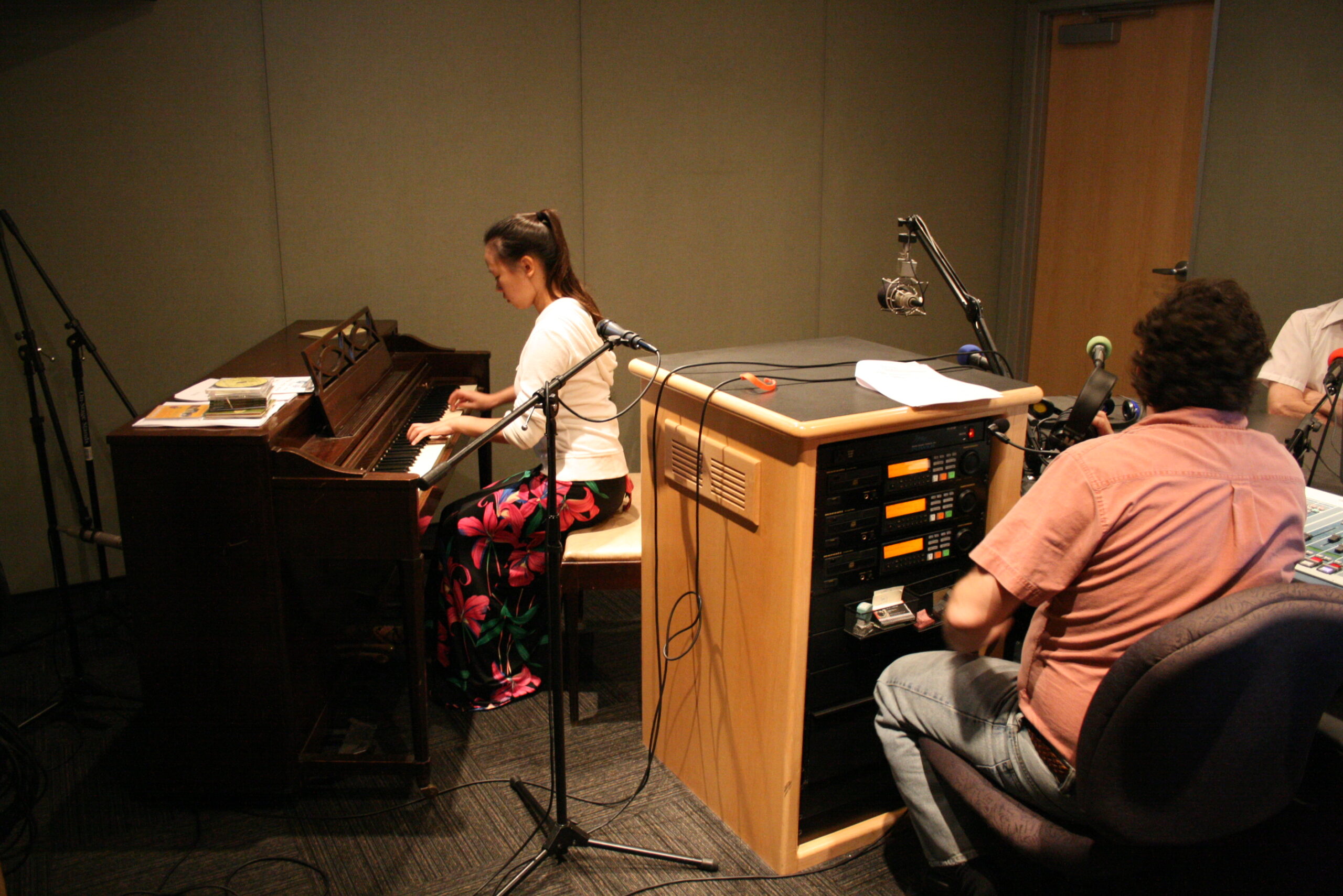 A teen playing a piano as an adult wearing headphones listens in a radio studio