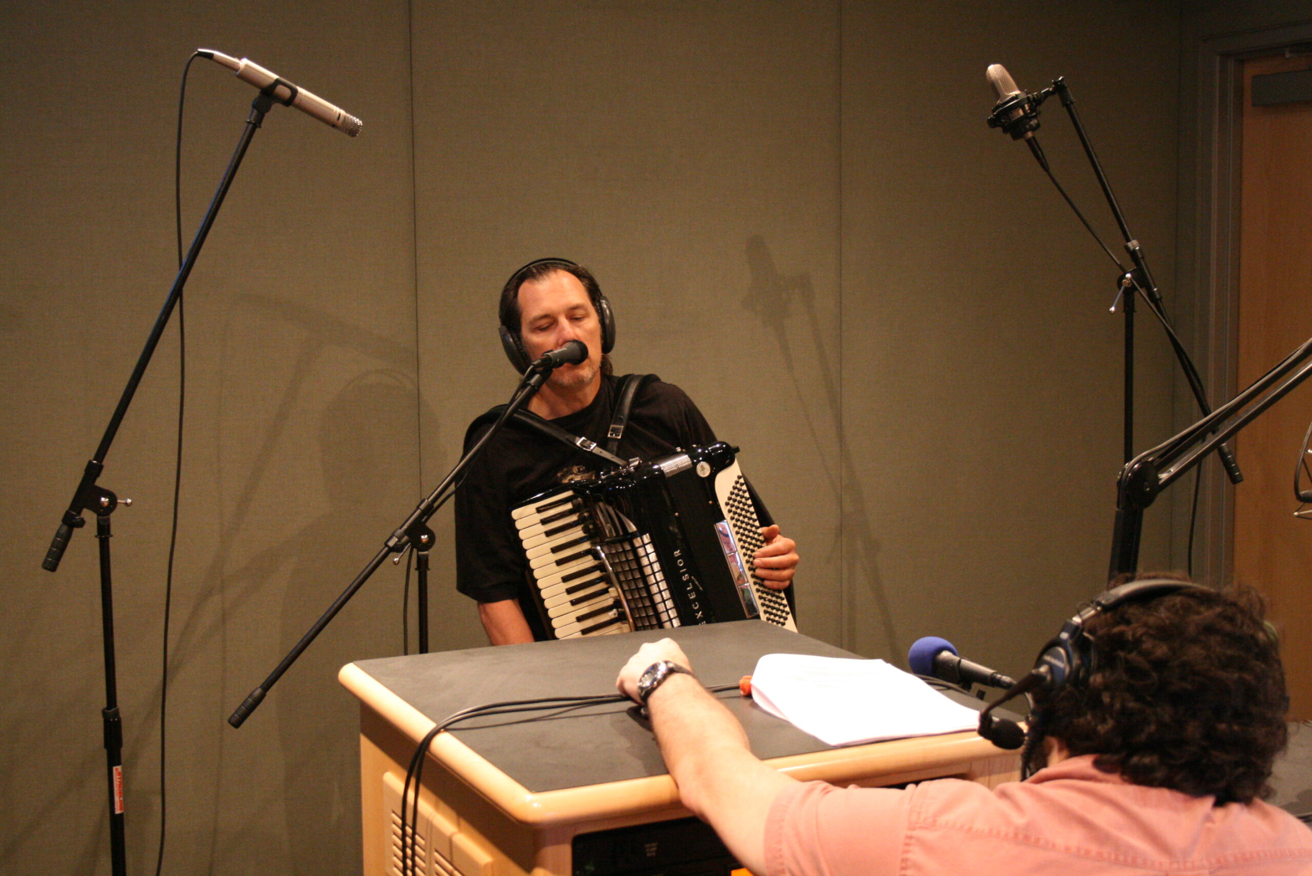 An adult playing an accordion as an adult wearing headphones listens in a radio studio