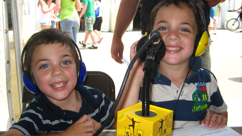 Two youth wearing headphones smiling while sitting behind a microphone