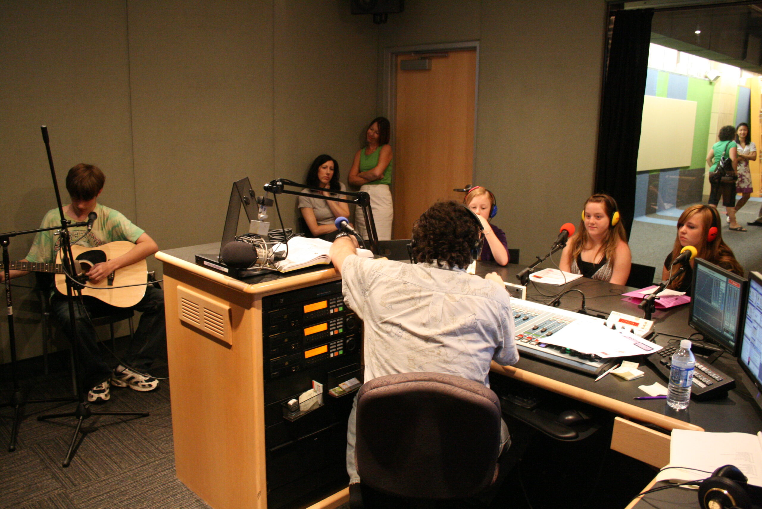 A youth playing an acoustic guitar, two adults in the background, and three youth wearing headphones sitting behind microphones being interviewed by an adult wearing headphones in a radio studio