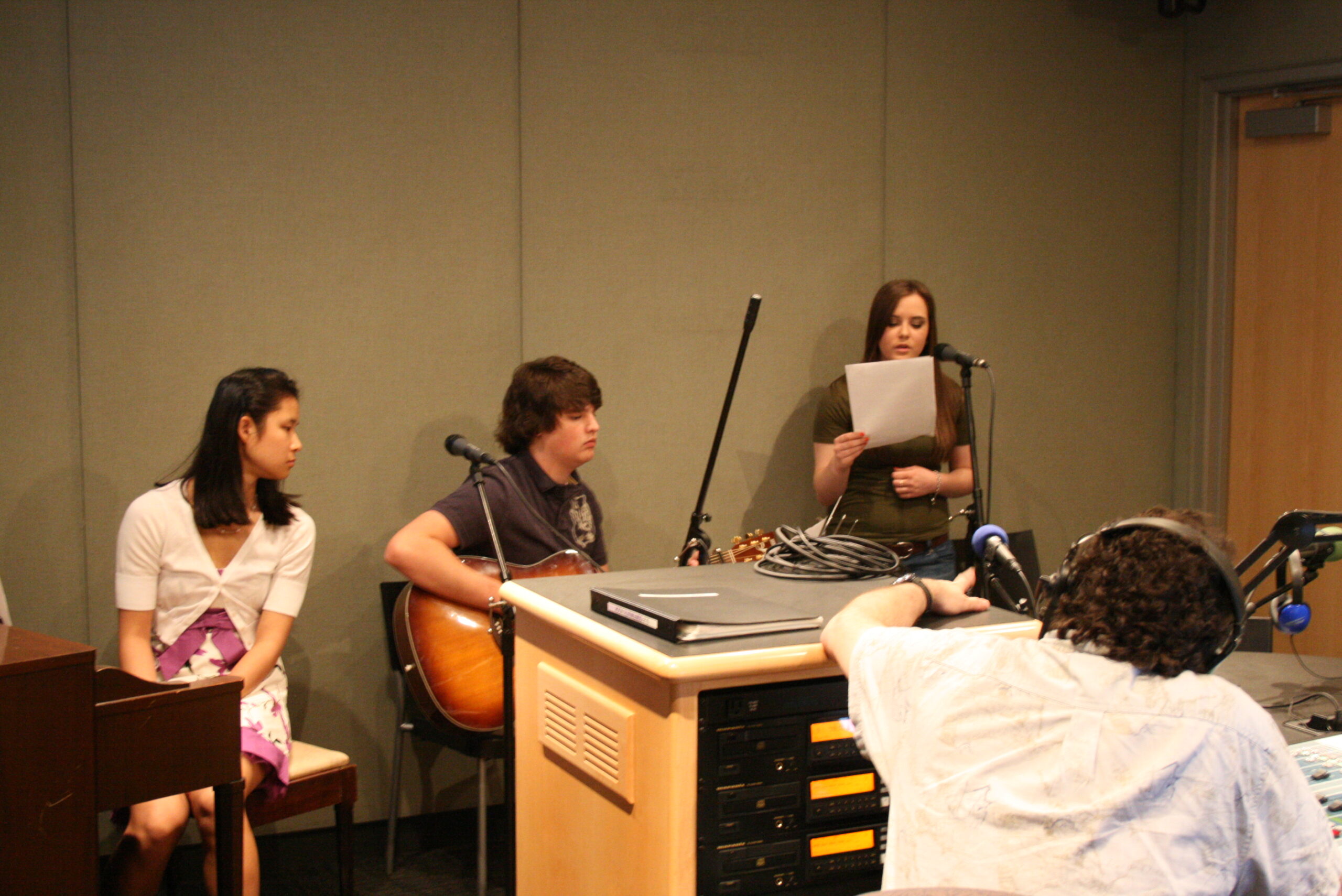 A teen sitting at a piano, a teen holding an acoustic guitar, and a teen holding a piece of paper and sitting behind a microphone being interviewed by an adult wearing headphones in a radio studio